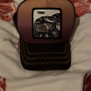 Flerfärgad keps med ormmotiv - Snygg keps med ett coolt ormmotiv och texten 'TOXIC' på framsidan. Kepsen har en gradient i orange och lila samt dekorativa sömmar på skärmen. Perfekt för en unik stil.