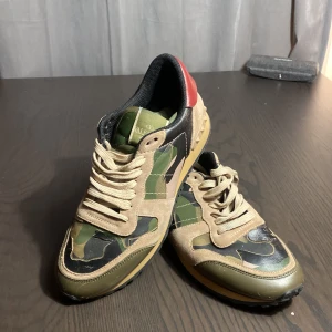 Valentino Rock Runners skor - Sneakers från Valentino i en snygg camouflage colorway i grönt, beige och svart. Skorna har en röd häl och är tillverkade i en kombination av skinn och mocka. De har snörning och en bekväm sula med nitar på baksidan. Lite använda
