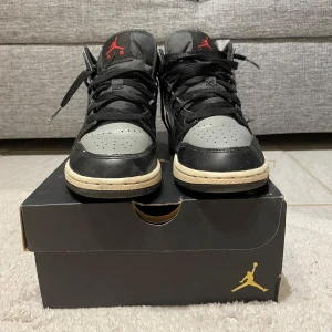 Nike Air Jordan 1 Mid (GS) i svart och grå - Säljer ett par Nike Air Jordan 1 Mid (GS) i svart och grå med röda detaljer. Skorna har en klassisk design med snörning och en bekväm passform. Perfekta för dig som älskar stilrena sneakers med en touch av färg.