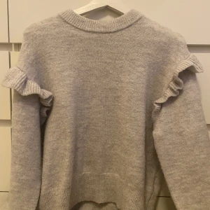 Grå stickad tröja med volangdetaljer - Säljer en mysig grå stickad tröja med volangdetaljer på axlarna. Tröjan har långa ärmar och en rund halsringning. Perfekt för kyliga dagar!