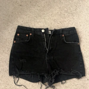Svarta jeansshorts från Zapa - Snygga svarta jeansshorts från Zapa med slitna detaljer och fransar. De har en dragkedja framtill och klassiska femficksdesign. Perfekta för en avslappnad stil.
