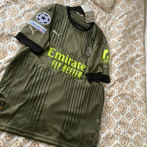 Olivgrön Ac Milan fotbollströja från Puma - Säljer en olivgrön fotbollströja från Puma med svarta detaljer och korta ärmar. Tröjan har tryck från Emirates och BitMEX samt UEFA Foundation-märke på ärmen. Perfekt för fotbollsfans som vill ha en unik look på planen. Samt att jag har aldrig använd den och har köpt den från hemsidan och såklart ÄKTA!. Priset sänks vid snabb affär. Köpte för 1099kr