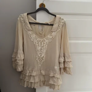 Beige blus från Odd Molly - Charmig beige blus från Odd Molly med vackra spetsdetaljer och volanger. Blusen har trekvartsärmar och en knytning i nacken 🕊️