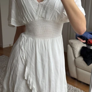 Studentklänning - Säljer en vacker vit klänning med smockdetalj i midjan och v-ringad hals. Klänningen har korta ärmar och ett luftigt fall, perfekt för varma dagar. Den har en omlott-look framtill som ger en extra touch. Perfekt för sommarens alla tillfällen såsom studenten! 