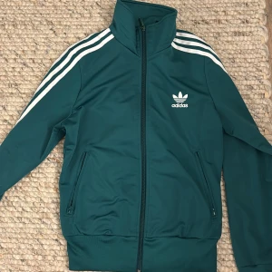 Grön träningsjacka från Adidas - Säljer en grön träningsjacka från Adidas med de klassiska tre vita ränderna längs ärmarna. Jackan har dragkedja framtill och två fickor med dragkedjor. Perfekt för en sportig look eller träning.