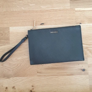 Svart kuvertväska från Mango - Snygg svart kuvertväska från Mango med en praktisk handledsrem och dragkedja i guld. Perfekt för att bära det viktigaste med stil. Enkel och elegant design som passar till många tillfällen. 21×14cm