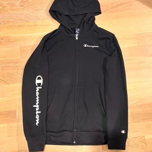 Svart hoodie från Champion - Säljer en svart hoodie från Champion med dragkedja och huva. Den har Champion-loggan tryckt på bröstet och ärmen. 