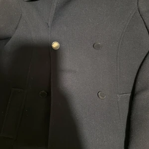 Mörkblå kappa från Holly & Whyte - Säljer en stilren mörkblå kappa från Holly & Whyte by Lindex. Kappan har en klassisk design med knappar framtill och två fickor. Perfekt för kyligare dagar och ger en elegant look.