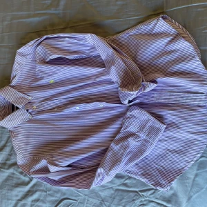 Lila och vit randig skjorta från Ralph Lauren - Säljer en stilren lila och vit randig skjorta från Ralph Lauren. Skjortan är i slim fit och har en broderad gul logga på bröstet. Tillverkad i 100% bomull, perfekt för en snygg och bekväm look. Knappar framtill och klassisk krage.