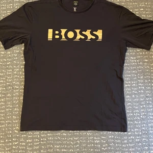 Svart t-shirt från BOSS - Snygg svart t-shirt från BOSS med ett stilrent guldtryck på bröstet. Tillverkad i mjuk bomull som stödjer Cotton made in Africa-initiativet. Perfekt för en avslappnad och trendig look.