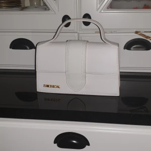 Vit handväska från Jacquemus - Snygg vit handväska från Jacquemus i stilren design. Väskan har ett handtag och en elegant stängning framtill. Perfekt för att ge en touch av lyx till din outfit. Den har en diskret ficka på baksidan för extra förvaring.