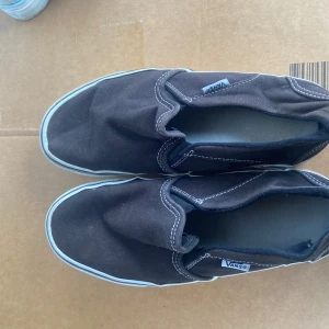 Svarta slip-on sneakers från Vans - Säljer ett par klassiska svarta slip-on sneakers från Vans. Skorna har en enkel design med vit sula och synliga sömmar. Perfekta för en avslappnad stil.