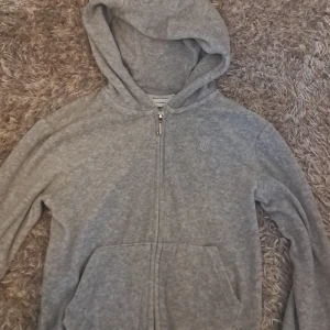 Grå hoodie från Juicy Couture - Säljer en stilren grå hoodie från Juicy Couture. Den är använd flera gånger men är i super bra skick. Det är ett litet fel som hände i fabriken när de gjorde tröjan men den är fortfrande supersnygg. Storleken är mellan 140-150.