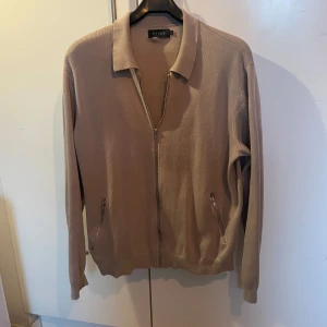 Beige tröja med dragkedja från Riley - Snygg beige tröja från Riley med dragkedja framtill och krage. Tröjan har långa ärmar och ribbade muddar vid ärmslut och nederkant. Perfekt för en stilren look.