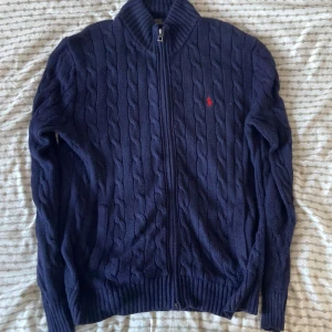 Mörkblå stickad kofta från Ralph Lauren - Säljer en snygg mörkblå stickad kofta från Ralph Lauren med dragkedja och kabelstickat mönster. Den har en broderad röd logotyp på bröstet och ribbade muddar vid ärmslut och nederkant. Perfekt för kyliga dagar!