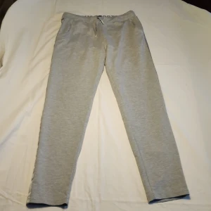 Grå mjukisbyxor från MM Sports - Säljer ett par bekväma grå mjukisbyxor från MM Sports. Byxorna har en elastisk midja med snörning för justerbar passform. Perfekta för en avslappnad dag hemma eller lätt träning.