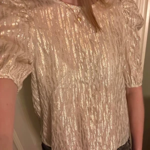 Glittrig topp från Holly & Whyte - Säljer en glittrig topp från Holly & Whyte by Lindex i storlek S. Toppen har korta puffärmar och ett glittrigt mönster i guld på en vit bas. Perfekt för att lysa upp vilken outfit som helst! ✨ Kan kännas lite sticksig eftersom det är glitter på insidan också, så jag rekommenderar att ha ett vitt linne under!