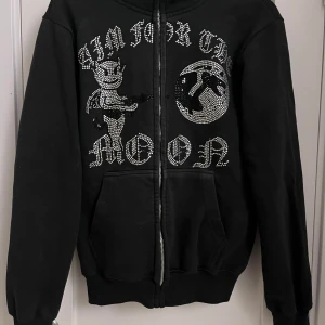 Svart hoodie med strassdetaljer - Säljer en svart hoodie med coola strassdetaljer och texten 'Aim for the Moon'. Den har en dragkedja framtill och långa ärmar. Perfekt för att sticka ut med en unik stil.