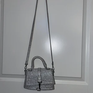 Glittrig silver axelväska - Snygg glittrig axelväska i silver med axelrem som går att ta av och handtag. Perfekt för att ge lite extra bling till din outfit. Lite fläckar på insidan men inget man tänker på alls (syns på sista bilden) 💕tror ej den säljs längre men vet ej 100 