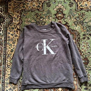 Grå sweatshirt från Calvin Klein Jeans - Säljer en stilren grå sweatshirt från Calvin Klein Jeans med det ikoniska CK-logotypen tryckt på framsidan. Tröjan har långa ärmar och en rund halsringning. Perfekt för en avslappnad look.
