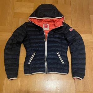 Mörkblå pufferjacka från Colmar - Säljer en blå pufferjacka från Colmar med en snygg orange insida. Jackan har en dragkedja framtill och två fickor med dragkedjor. Den har en huva och är perfekt för kyligare dagar. Märkeslogga på ärmen ger en extra touch. Passar perfekt nu till våren