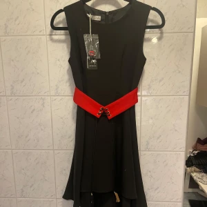 Svart klänning med rött bälte - Elegant svart klänning med ett iögonfallande rött bälte med snörning framtill. Klänningen är ärmlös och har en dragkedja i ryggen för enkel påklädning. Perfekt för en stilren look.