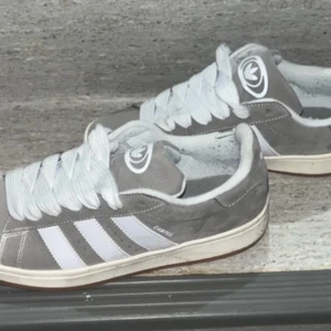 Grå Adidas Campus sneakers - Snygga grå Adidas Campus sneakers i mocka med vita detaljer och snörning. De har den klassiska tre-randiga designen på sidorna och Adidas-loggan på plösen och hälen. Perfekta för en stilren och avslappnad look.