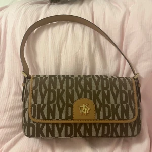 Brun axelväska från DKNY - Snygg brun axelväska från DKNY. Väskan har en justerbar axelrem och ett magnetlås. Insidan är rymlig med en dragkedjeficka för extra säkerhet. Små missfärgningar på loggan och guldspännerna. Nypris ca 2300kr