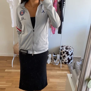 Grå träningsjacka från Jordan x PSG - Säljer en grå träningsjacka från Jordan x PSG med dragkedja och långa ärmar. Jackan har en logga på bröstet och röd-vit-blå randdetalj på ärmen. Perfekt för sportiga tillfällen eller som en stilren vardagsjacka. Den är i barn storlek men passar mig som har Xs
