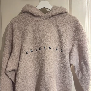 Beige fleece hoodie från Jack & Jones - Mysig beige hoodie från Jack & Jones Originals i fleece-material. Perfekt för kyliga dagar med sin varma och bekväma design. Den har en luva och texten 'ORIGINALS' tryckt på framsidan. Nypris 400