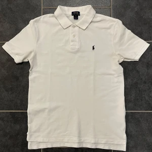 Vit pikétröja från Ralph Lauren - Säljer en klassisk vit pikétröja från Ralph Lauren med den ikoniska loggan broderad på bröstet. Tröjan har korta ärmar och en traditionell krage med knappar. Perfekt för en stilren look. Storlek ”L” men sitter som en S