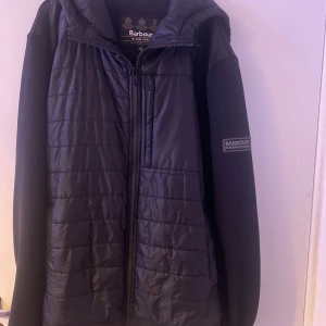 Svart jacka från Barbour International - Säljer en svart jacka från Barbour International med dragkedja och huva. Jackan har en quiltad framsida och är långärmad. Perfekt för kyligare dagar. Passar även för dom som har storlek L 