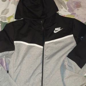 Grå och svart huvtröja från Nike - Säljer en snygg grå och svart huvtröja från Nike med dragkedja framtill. Tröjan har en huva och långa ärmar, perfekt för en sportig look. Den har även en vit randdetalj över bröstet och Nike-loggan på bröstet.