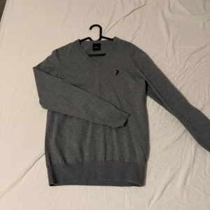 Grå v-ringad tröja från Ralph Lauren - Snygg grå v-ringad tröja från Ralph Lauren med broderad logga på bröstet. Tröjan har långa ärmar och ribbade muddar vid ärmslut och nederkant. Perfekt för en stilren look. Stolen M men passar även som storlek S.