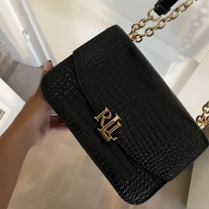 Ralph Lauren väska - Handväska från Lauren Ralph Lauren. Modellen är Madison.  svart med gulddetaljer  och guldkedja Går göra långt band eller kort  Två fack inne i väskan, ett fack med dragkedja. Ett fack utanpå baksidan, mobilfack.  Mått ca: 23*16*7  Nypris 4000kr Skickas s