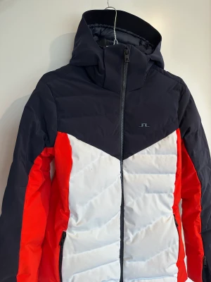 J. Lindeberg Crystal down ski jacket Rojo skidjacka - Designad för maximal komfort, kombinerat m slitstarkt vattentätt skydd. En anmärkning är på höger bröst på den vita delen upptill, se bild, där den fått sig ett svagt blå svart knappt synbar märke. Utrustad med precis allt man behöver i en varm skidjacka.