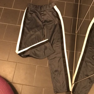 Träningsbyxor - Träningsbyxor i glansigt material från Boohoo. Mörkgrå med svarta och vita revär och inga fickor. Nya med lappen kvarstorlek 36. 