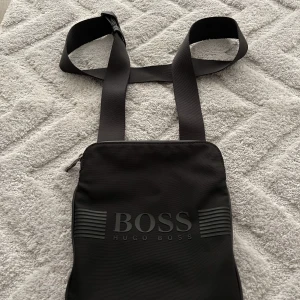 Hugo boss axelväska - Jag köpte den för 1100 kr i början på sommarn och jag använde axelväskan bara på sommaren. Den är fortfarande i ett väldigt fint skick. Man kan ha plats med många saker som till exempel plånbok och telefon. Den är väldigt skön och lätt att ha med sig. 