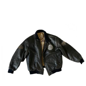 Jacka  - BRUNGRÅ JACKA I ÄTKA LÄDER MED FLERA FETA BADGES                                                  SKICK: Jackan är i bra skick och har inga defekter. 