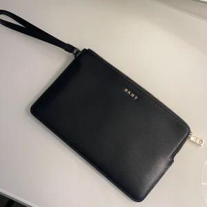 DKNY Pouch - Äkta DKNY BRYANT wrislet wallet pouch i svart. Helt oanvänd, köpt för ca 9 månader sedan på Zalando, säljer på grund av ingen användning. Bevis på köpet samt fler bilder skickas vid intresse. Inget slitage. Köpt för 929 kronor. Mottagaren betalar frakt. 