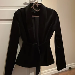 Svart kavaj - Kavaj i sammet. Jättefin och som jag fått komplimang för. Använd 2-3 ggr, blev sedan för liten. Knäppes med en liten hyska och det medföljer band ;också i sammet) som går att knyta om så önskas. Inga fickor. Ser helt oanvänd ut. Nypris 499 på Vila