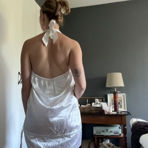 Satinklämning med öppen rygg i pärlemovit  - Superskön Santin klänning i polyester. Den har varit med om födelsedagar, nyår och andra fester. Lätt att dra på sig och genast se festlig ut. Knytning i nacken men öppen rygg, snörning vid hem för det möjligt att själv välja hur klänning ska sitta. Pärl