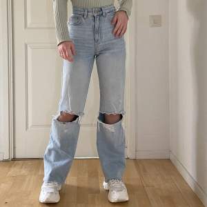 Ett par snygga ljusblåa jeans med slitna knä. De är högmidjade och vida i benen. Jeansen är klippta i längden så de passar korta/petite tjejer. Jeansen är använda och det finns en liten fläck på höger ben längst ner, men de är fortfarande i bra skick.