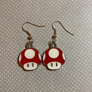 Super Mario Örhängen Röd - Nya Super Mario inspirerade örhängen, perfekt till Nintendo älskaren! Svampen är ca 1,6cm som störst. Föredrar Swish, skickar bara och möts ej upp helst.