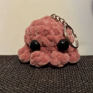 Virkad minibläckfisk-nyckelring - Egengjord, virkad minibläckfisk med nyckelring, i nyskick. Garnet är 100% polyester. Den är ca 6 cm lång och 5 cm hög. Den kostar 39kr totalt med frakt!
