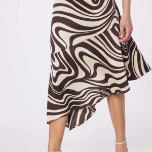 Kjol  - Storlek 40 A-linje-kjol med volang och assymetrisk fåll i satin. Dragkedja i sidan och med bruna prins på ljus botten. Sparsamt använd, inga anmärkningar. Pris 200 kr 