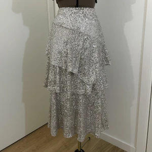 Silvrig kjol i paljetter från Topshop  - Engångsanvänd volangkjol i midilängd med silvriga paljetter och resårmidja. Materialet är strechigt  Längd: ca 80cm