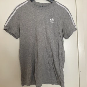 Grå Adidas t-shirt - Väl använd t-shirt från Adidas. Den är fin och har ingen direkt defekter, fast materialet är lite ”slitent” efter att ha varit använd och tvättat et antal gånger. Relaxed fit. Skriv om det önskas fler bilder eller information 🌸