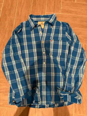 Blå rutig skjorta från Hollister - Säljer en blå och vit rutig skjorta från Hollister i storlek L. Skjortan har klassisk krage, långa ärmar och knäppning framtill. Passar mer för storlek M. Perfekt för dig som gillar en avslappnad men ändå stilren look. Materialet är mjukt och skönt bomull.