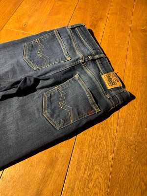 Mörkblå smala jeans med kontrastsömmar - Snygga mörkblå jeans med smal passform och kontrastsömmar. Jeansen har klassiska fem fickor och coola detaljer på bakfickorna. Materialet är mjukt denim med lätt stretch för extra komfort. Perfekta för dig som gillar en clean och stilren look lappen där det står replay där bak är lite av riven.